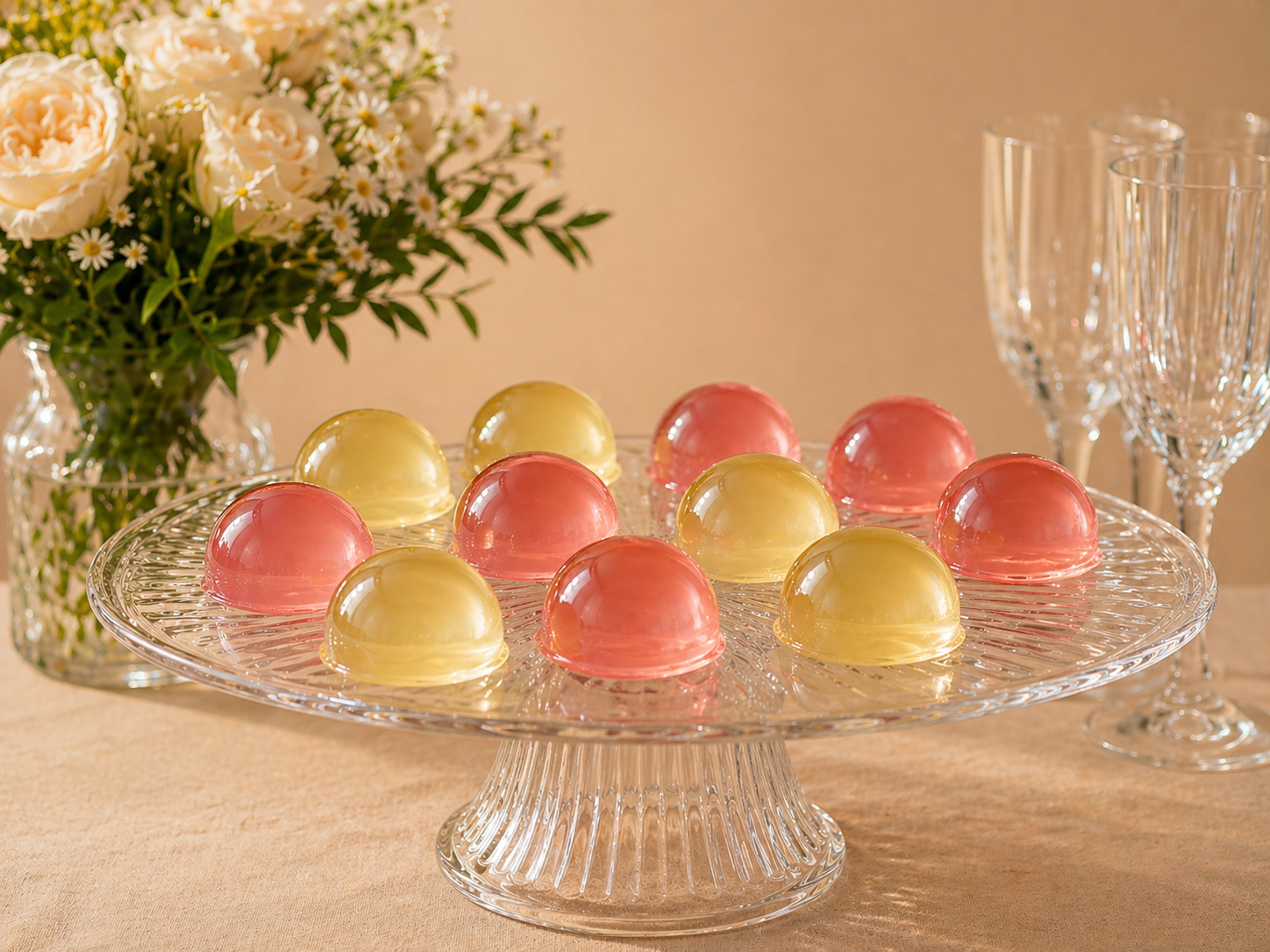 Luxury glass pedestal display of smooth dome jellies in blush pink and golden yellow tones, styled with flowers and champagne flutes for celebrations and events.